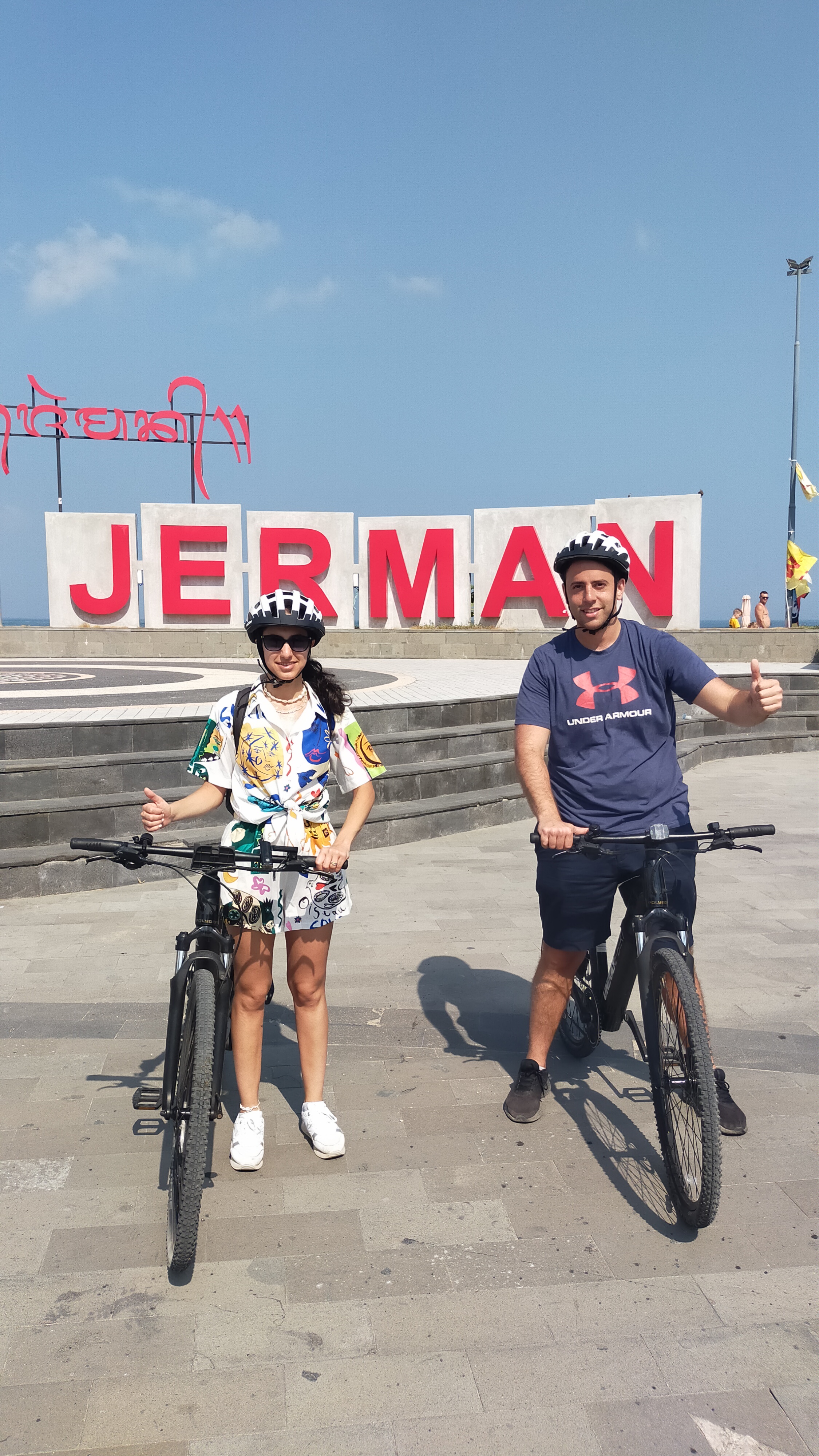 Two happy UNIKAT E-Bike riders posing with thumbs up at the Jerman sign on Pantai Jerman beach in Bali